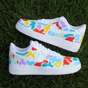May include: White low-top sneakers with a colorful camouflage pattern. The pattern includes shades of red, yellow, blue, green, and purple. The sneakers have a white sole and a white swoosh logo on the side.