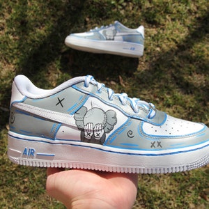 K&ouml;nnte beinhalten: Wei&szlig;e Low-Top-Sneaker mit blauen Akzenten und einem grauen und wei&szlig;en Grafik eines Charakters mit einem 'X' auf der Stirn und der sich die Augen mit den H&auml;nden bedeckt. Der Text 'AIR' befindet sich an der Seite des Schuhs.