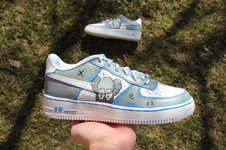 K&ouml;nnte beinhalten: Wei&szlig;e Low-Top-Sneaker mit blauen Akzenten und einem grauen und wei&szlig;en Grafik eines Charakters mit einem 'X' auf der Stirn und der sich die Augen mit den H&auml;nden bedeckt. Der Text 'AIR' befindet sich an der Seite des Schuhs.