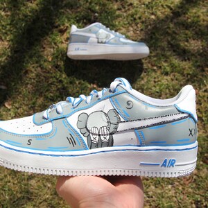 K&ouml;nnte beinhalten: Individuelle gestaltete wei&szlig;e und graue Sneaker mit blauen Akzenten. Die Sneaker zeigen eine Comic-Zeichnung eines Charakters mit den H&auml;nden vor dem Gesicht. Der Text "AIR" ist auf der Sohle des Schuhs sichtbar.