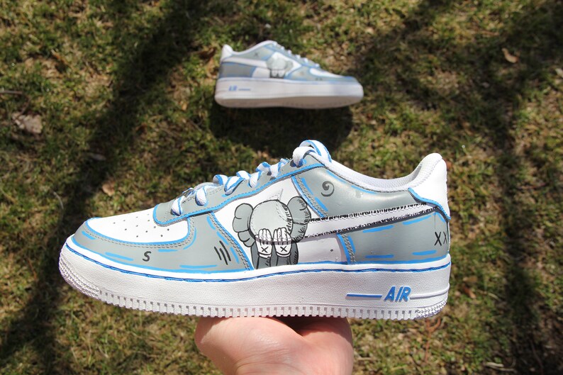 K&ouml;nnte beinhalten: Individuelle gestaltete wei&szlig;e und graue Sneaker mit blauen Akzenten. Die Sneaker zeigen eine Comic-Zeichnung eines Charakters mit den H&auml;nden vor dem Gesicht. Der Text "AIR" ist auf der Sohle des Schuhs sichtbar.