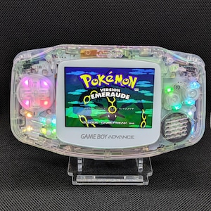 Puede incluir: Una consola portátil Game Boy Advance transparente con el juego Pokémon Esmeralda en la pantalla. La consola tiene una carcasa transparente, que revela los componentes internos y luces LED de colores. La pantalla muestra el logotipo del juego Pokémon Esmeralda.