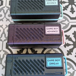 Puede incluir: Tres fundas para consolas de juegos portátiles en negro, morado y azul. Cada funda tiene una etiqueta rectangular que dice "GAME BOY ADVANCE" o "GAME BOY DMG-01". Las fundas tienen líneas diagonales y asas de colores.