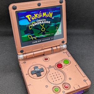 Könnte beinhalten: Kupferfarbene Game Boy Advance SP-Konsole, geöffnet, um das Spiel "Pokémon Version Émeraude" anzuzeigen. Die Konsole verfügt über ein Steuerkreuz, rote und grüne Knöpfe und den Text "GAME BOY ADVANCE SP".
