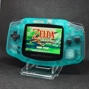 Könnte beinhalten: Ein türkisgrüner Game Boy Advance Handheld-Konsolen mit dem Spiel "The Legend of Zelda: The Minish Cap". Die Konsole hat ein transparentes Gehäuse, schwarze Knöpfe und einen transparenten Kunststoffständer. Der Bildschirm zeigt eine lebendige Spielszene.