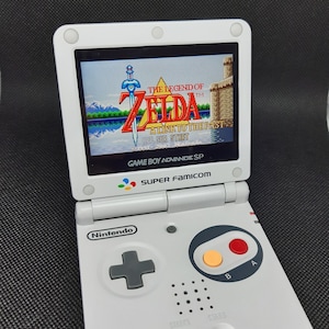 Peut inclure: Une console de jeu portable Game Boy Advance SP blanche avec un écran noir et blanc affichant l'écran titre du jeu "The Legend of Zelda: A Link to the Past". La console a un logo Nintendo gris, un pavé directionnel gris et des boutons gris étiquetés "A", "B", "Start" et "Select".