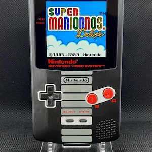 Op de afbeelding: Een zwarte draagbare videogameconsole met een scherm dat het titelscherm van Super Mario Bros. Deluxe weergeeft. Het apparaat heeft een richtingspad, twee rode A- en B-knoppen, en Select- en Start-knoppen, met Nintendo-branding op de voorzijde.