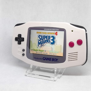 Peut inclure: Console portable Nintendo Game Boy Advance blanche, affichant Super Mario Bros. 3. La console a une croix directionnelle noire, des boutons roses et le logo Nintendo. L'écran affiche le titre et le gameplay du jeu.