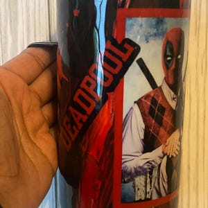 May include: A black and red stainless steel tumbler featuring a Deadpool image and the word "DEADPOOL" in white lettering. The tumbler has a black lid and a black handle.