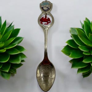 May include: A silver souvenir spoon with a red crab charm and a shield-shaped top with a colorful design. The spoon bowl features an engraved image of a ship and text. A decorative collectible.