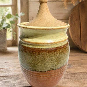 May include: A ceramic jar with a conical lid, featuring a multi-toned glaze. The base is a warm terracotta orange, transitioning into a mossy green midsection, and topped with a pale, creamy yellow rim and a textured, tan-colored lid.