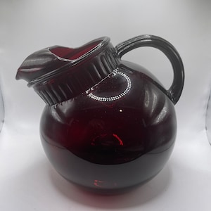 May include: A dark red glass pitcher with a round body and a handle. The pitcher has a textured rim and a glossy finish.