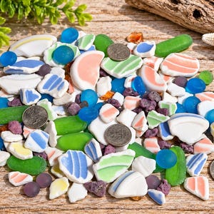 May include: A collection of multi-colored sea glass and ceramic shards in shades of blue, green, white, pink, and yellow, interspersed with small purple pebbles and three silver United States quarters for scale on a weathered wooden surface.