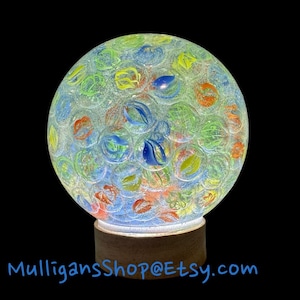 May include: A clear glass paperweight with a wooden base. The paperweight is filled with colorful marbles, including blue, green, yellow, and orange.