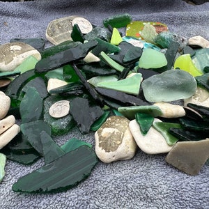 May include: A collection of sea glass in various shades of green, including some clear and white pieces. The glass is smooth and rounded from being tumbled by the ocean.