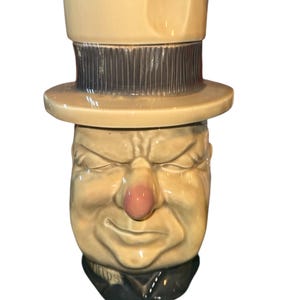 May include: A vintage ceramic pitcher shaped like a man's face wearing a top hat. The face is tan with a pink nose and closed eyes. The hat is tan with a dark gray band. The base of the pitcher has the word "FIELDS" on it.