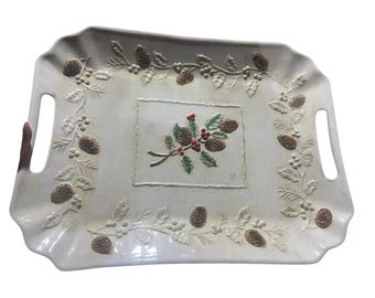 May include: Cream-colored rectangular serving tray with embossed pinecones, holly, and berries. The tray has two handles and a central rectangular design with a pinecone and berry motif. The tray's edges are scalloped.