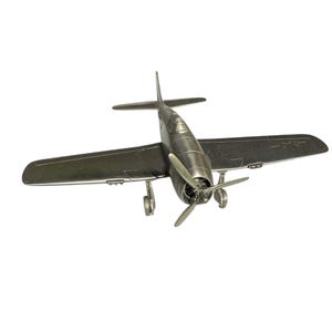 May include: A detailed metal model airplane with a propeller and wings. The aircraft is silver and has a classic design, with visible landing gear and a cockpit. The model is set against a white background.