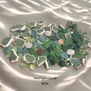 May include: A collection of green and white sea glass and ceramic pieces. The pieces are arranged on a white surface. There are also a few coins scattered among the sea glass.