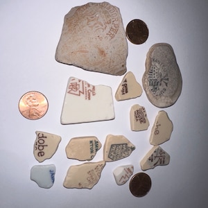 May include: A collection of vintage ceramic tile fragments with various designs and text, including "Made in China", "Mell's", "Ho-Toin", "Acuse Mina", "Jacks", "Amnton", "Syrac", "dobe", and "he". There are also two copper-colored coins.