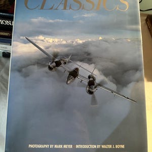 May include: A vintage book titled "Classics" with a photograph of a silver airplane flying through clouds. The book cover features the text "Photography by Mark Meyer Introduction by Walter J. Boyne".