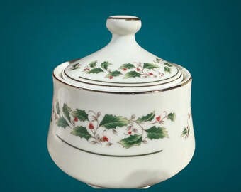 May include: A white ceramic sugar bowl with a lid, adorned with a holly and berry design. The bowl has a gold trim and a small knob on the lid. The holly leaves are green, and the berries are red.
