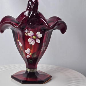 May include: A deep red glass basket with a ruffled rim and a high arched handle. The body features hand-painted white and purple flowers with green leaves and a small bee. The base is hexagonal and includes a thin silver-colored band near the stem.