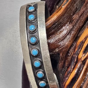 May include: A silver cuff bracelet featuring a central dark oxidized channel inlaid with eight small, round, bright blue turquoise stones. The metal band has a smooth, polished finish on the outer edges, framing the row of stones.