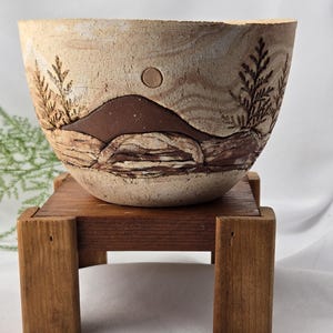 May include: A ceramic bowl with a textured, light beige surface featuring a dark brown mountain silhouette, a circular sun, and etched pine tree designs. The bowl has a marbled appearance with horizontal layers and sits on a small wooden stand.