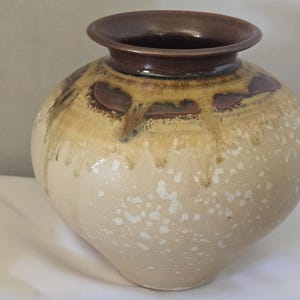 Puede incluir: Un jarrón de cerámica redondo con una abertura ancha y un borde marrón oscuro. El cuerpo presenta una base de color crema con patrones de manchas blancas y un efecto de esmalte goteado en tonos bronce y marrón oscuro desde el cuello.