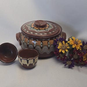 May include: A brown ceramic pot with a lid and two side handles, accompanied by two matching small bowls. The set features a decorative pattern in white, black, and yellow. A cluster of artificial yellow and purple flowers sits to the right.