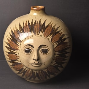 Puede incluir: Un jarrón de cerámica redondo con un acabado beige brillante que presenta un diseño de cara de sol pintado a mano. El sol tiene rayos de color marrón oscuro que irradian hacia afuera, ojos detallados, nariz y boca.