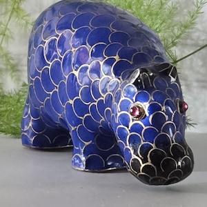 May include: A blue animal figurine featuring a repeating scale pattern outlined in gold. The creature has a rounded body, a snout with black detailing, and two small, circular, dark red gemstone eyes set into the head, creating a distinct visual contrast.