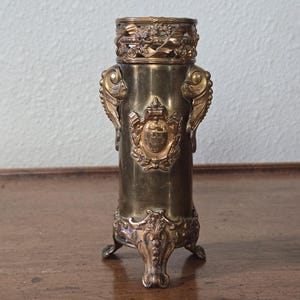 Op de afbeelding: Een cilindrische koperen vaas met een sierlijke opengewerkte rand met bloemmotieven, twee zijhandvatten in de vorm van gestileerde vissen en een centraal reliëfschild. De basis wordt ondersteund door drie decoratieve pootjes met krulwerk.