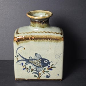 Peut inclure: Un vase en céramique de forme carrée avec un vernis crème clair et un bord brun. La face avant présente un motif de poisson bleu peint à la main entouré de petits accents floraux verts et bleus. Le vase a un col cylindrique court et une finition mouchetée.