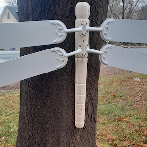 May include: A white dragonfly wind spinner with wooden body and four blades. The spinner is attached to a tree trunk.