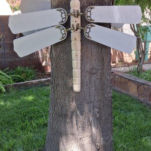 May include: A whimsical dragonfly sculpture made from repurposed ceiling fan blades, mounted on a tree trunk. The dragonfly features white blades and a wooden body, with decorative brass-colored accents. The sculpture is an outdoor garden art piece.