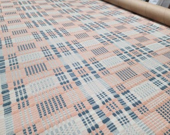 May include: A woven fabric featuring a repeating plaid pattern in soft peach, muted blue, and off-white. The textile displays a textured grid design with alternating blocks of color and vertical and horizontal lines throughout the material.
