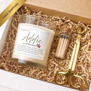 May include: A gift set containing a clear glass candle with a white label reading The Aroma of Addie A Loving Aunt, a small glass vial filled with wooden matches with a cork stopper, and a gold-toned metal wick trimmer inside a cardboard box.
