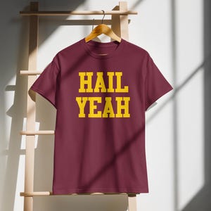 Pode incluir: Camiseta vinho com as palavras "HAIL YEAH" impressas em amarelo.