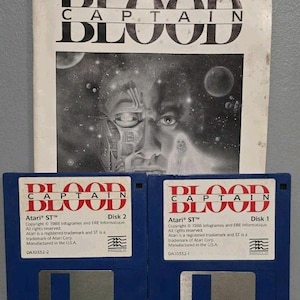 May include: A vintage Captain Blood video game set for Atari ST, featuring a paper manual with a black and white space-themed illustration and two blue 3.5-inch floppy disks labeled Disk 1 and Disk 2, each with white labels and red text.