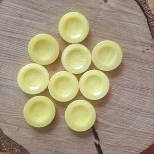 Może przedstawiać: Dziewięć żółtych, okrągłych guzików z wypukłym środkiem i subtelnym wzorem w paski. Guziki są ułożone w kształt gwiazdy na drewnianej powierzchni.