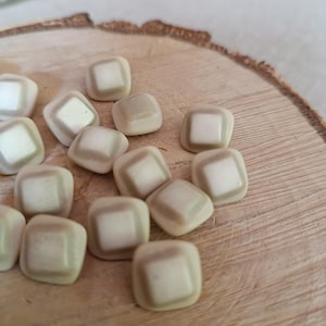 Może przedstawiać: Kolekcja kwadratowych, kremowych koralików z jasnobrązową obwódką. Koraliki są rozrzucone na drewnianej powierzchni, prezentując zaokrąglone rogi i subtelne wariacje kolorystyczne. Idealne do tworzenia biżuterii lub projektów rzemieślniczych.