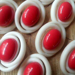 Può includere: Primo piano di diversi bottoni rossi e bianchi. I bottoni sono di forma ovale con un anello bianco attorno al centro rosso. I bottoni sono disposti a formare un motivo su una superficie di legno.