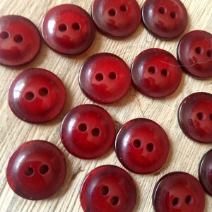 Puede incluir: Una colección de 15 botones de plástico rojo, cada uno con dos agujeros. Los botones están dispuestos en forma de semicírculo sobre una superficie de madera.