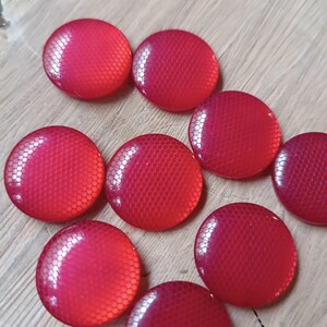 May include: A collection of eight red, round buttons with a textured, honeycomb-like pattern. The buttons are arranged on a light-colored wooden surface, showcasing their glossy finish and vibrant color.