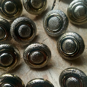 Może przedstawiać: Kolekcja 14 złotych guzików z wypukłym, fakturowanym środkiem i wzorem koncentrycznych kół. Guziki są ułożone w przypadkowy wzór na drewnianej powierzchni.