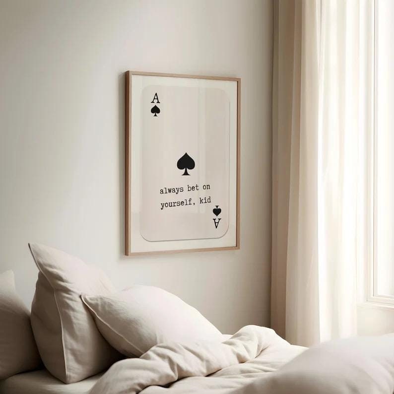 Puede incluir: Una impresi&oacute;n enmarcada de una carta de juego de as de picas con el texto "always bet on yourself, kid" impreso en ella. La impresi&oacute;n est&aacute; colgada en una pared blanca sobre una cama con ropa de cama blanca.
