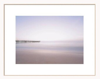 May include: A long wooden pier extends into a calm, pale blue ocean with a soft, pink-hued sky in the background.