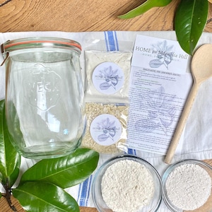 May include: A sourdough starter kit with instructions, a glass jar, a bag of flour, a bag of sourdough starter, and two bowls of flour. The kit is labelled "Home on Magnolia Hill".